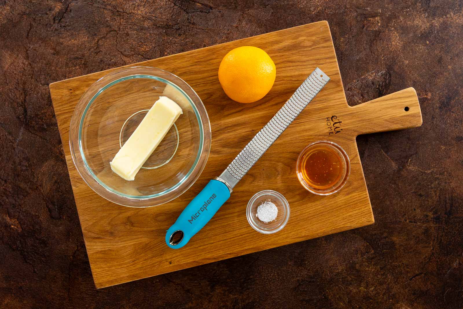 Ingredients for Orange Honey Butter arranged on a wooden cutting board: a stick of butter in a glass bowl, an orange, a Microplane zester, a small bowl of honey, and a small bowl of salt.