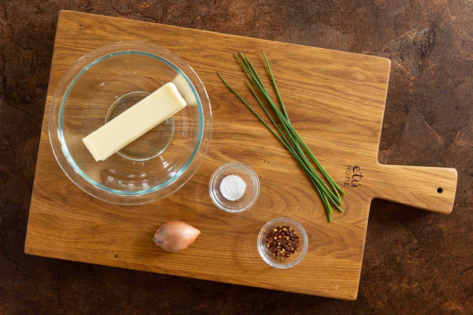 Ingredients for Que Sera Butter arranged on a wooden cutting board: a stick of butter in a glass bowl, fresh chives, a shallot, a small bowl of salt, and a small bowl of crushed red pepper flakes.