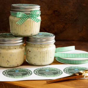 Small jars of savory Que Sera Butter tied with green gingham ribbon and topped with printed labels, arranged on a wooden board with scissors, ribbon, and label sheets.