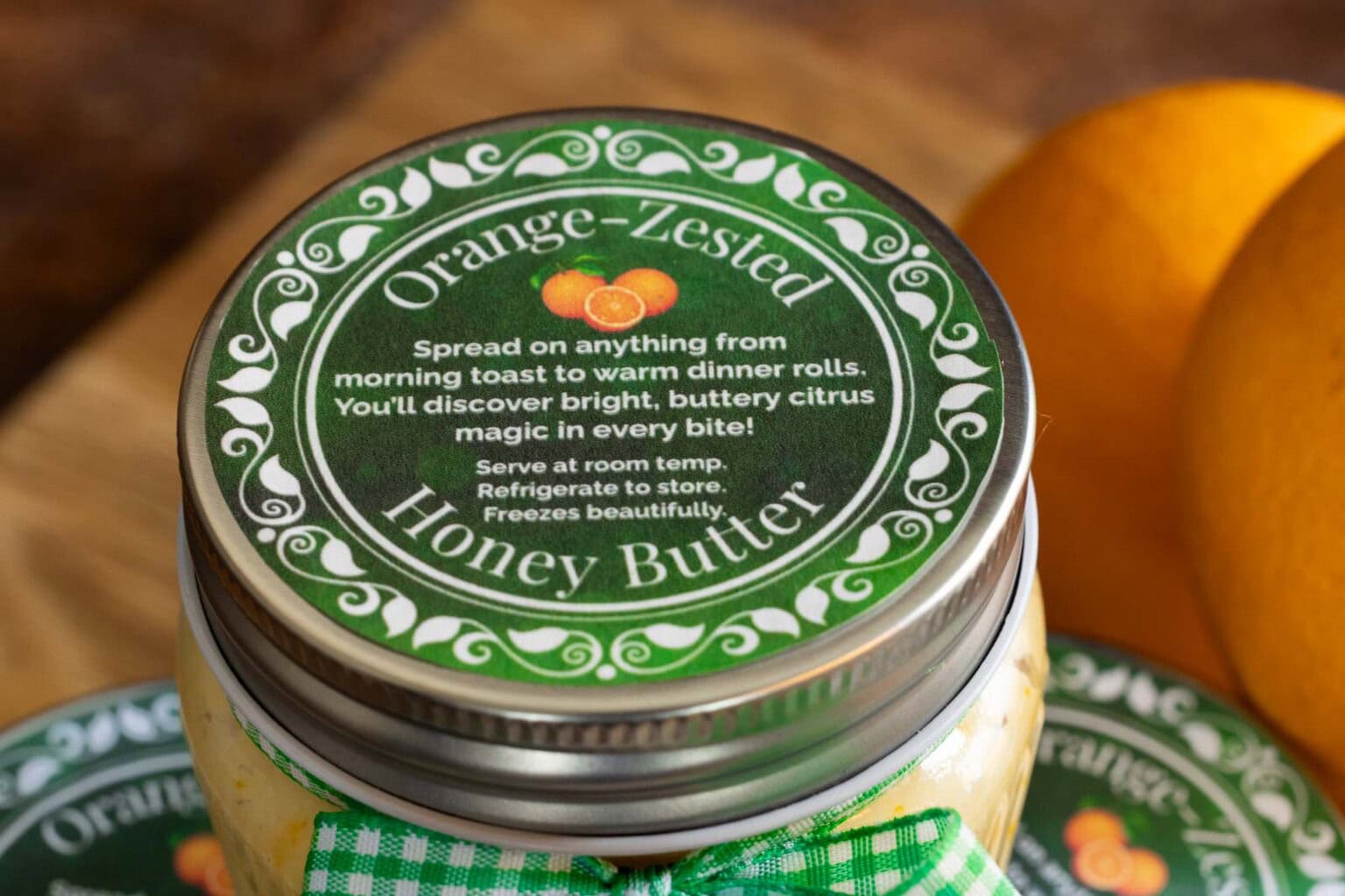 Close-up of a jar of Orange-Zested Honey Butter topped with a green printable label and tied with green gingham ribbon, with oranges and additional labeled jars in the background.