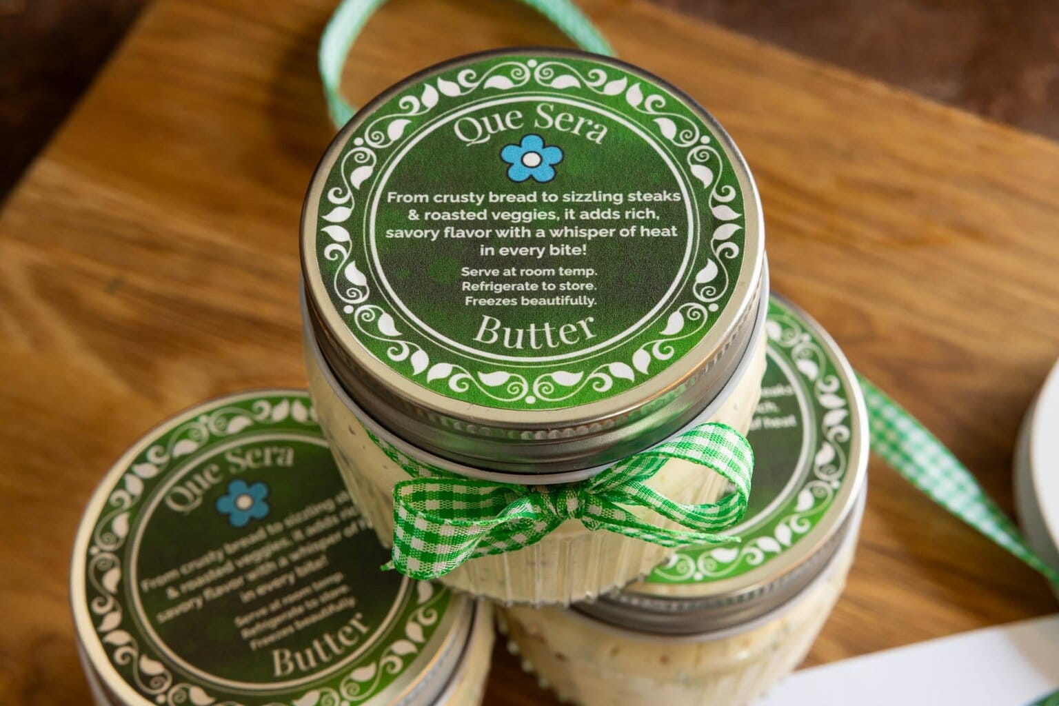 Close-up of a jar of Que Sera Butter topped with a green printable label and tied with a gingham ribbon, with additional jars and ribbon in the background.