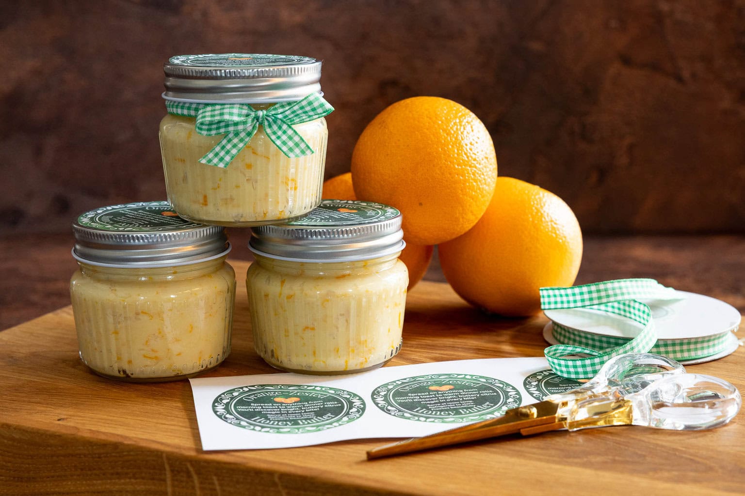 Jars of Orange Honey Butter tied with green gingham ribbon and topped with printable labels, arranged on a wooden board with fresh oranges, ribbon spools, scissors, and label sheets.