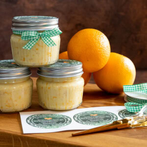 Small jars of Orange Honey Butter topped with gingham ribbon and green printable labels, displayed on a wooden board with fresh oranges, scissors, and ribbons in the background.