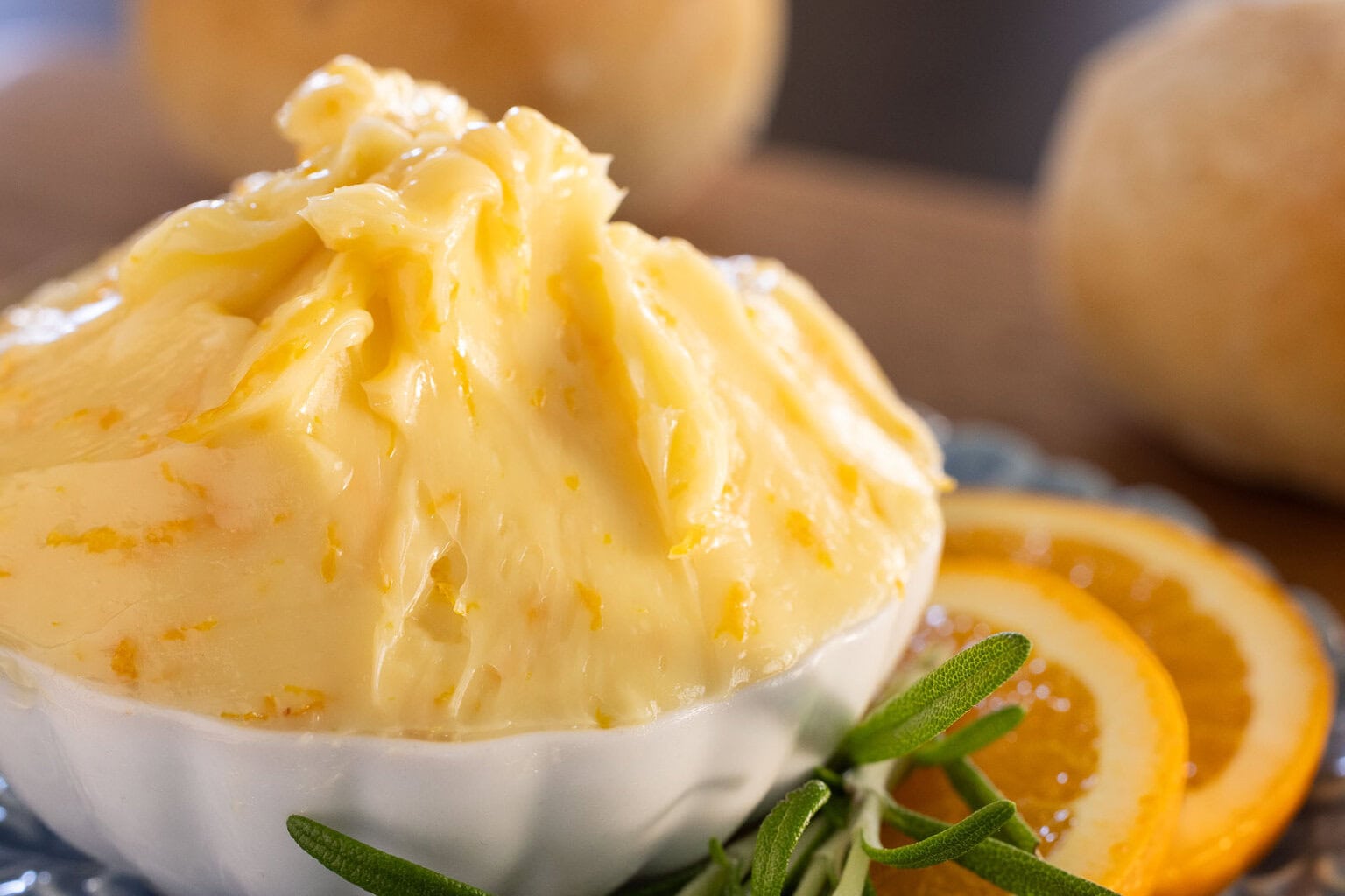 Close-up of creamy Orange Honey Butter in a white dish, flecked with orange zest, with fresh orange slices and rosemary beside it.