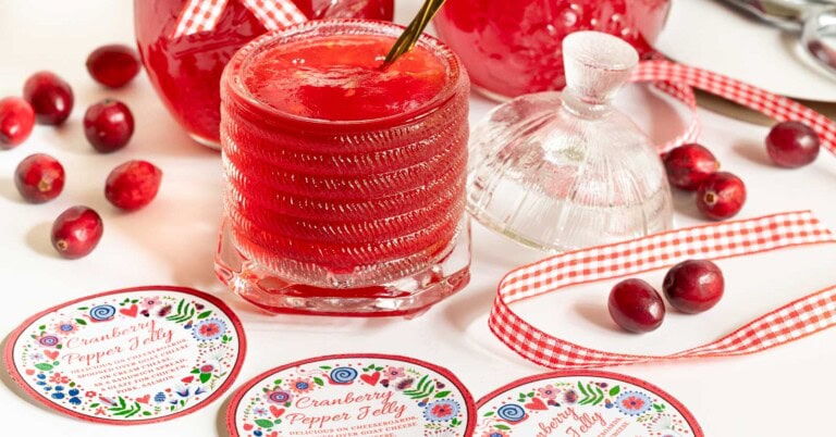 Glass jars of cranberry pepper jelly with cranberries, gingham ribbon, and round printable labels styled for holiday gifting