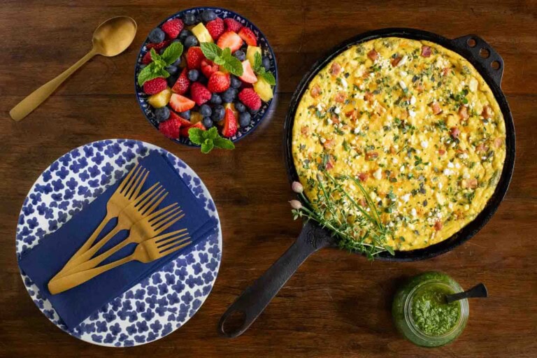 Fresh herb, ham and goat cheese frittata baked in a cast iron skillet, served with a bowl of mixed berries and a small jar of green herb sauce on a wooden table.