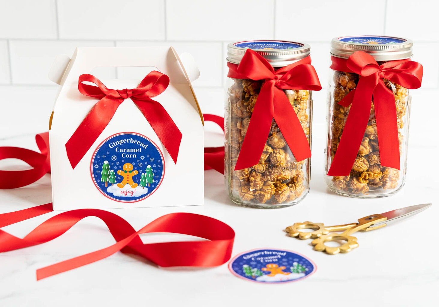 Jars and a small white gift box filled with Gingerbread Caramel Corn, each decorated with bright red ribbons and holiday labels, displayed with loose ribbon and scissors on a white surface.