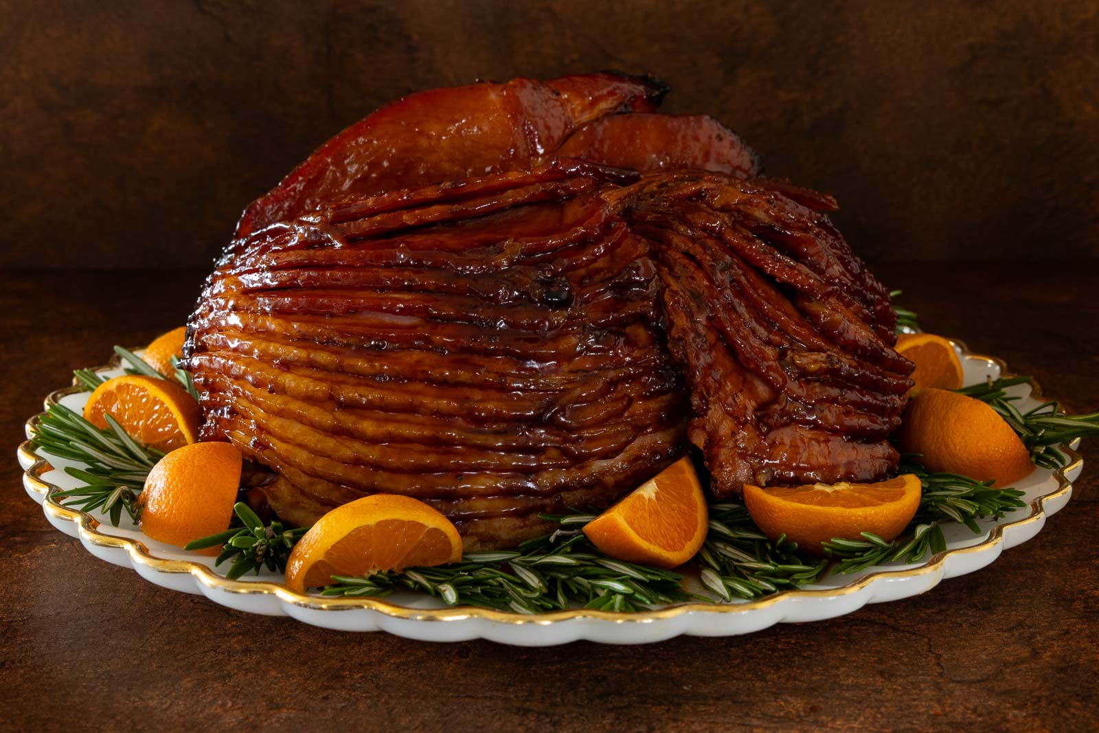 Glazed spiral ham on a serving platter, garnished with fresh rosemary and orange slices, set on a rustic wooden surface.