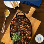 Overhead view of a serving dish filled with beef bourguignon-style stew with carrots, pearl onions, mushrooms, and rosemary on a wooden board, surrounded by gold flatware, blue napkins, a gold spoon, and small blue-and-white dishes.