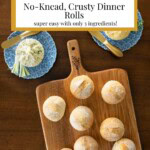 Overhead view of a wooden board filled with golden, crusty no-knead dinner rolls, surrounded by small blue plates with compound butters and jam on a wooden table.