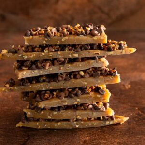 Stack of homemade English toffee pieces topped with chocolate chips and toasted pecans on a rustic brown background.