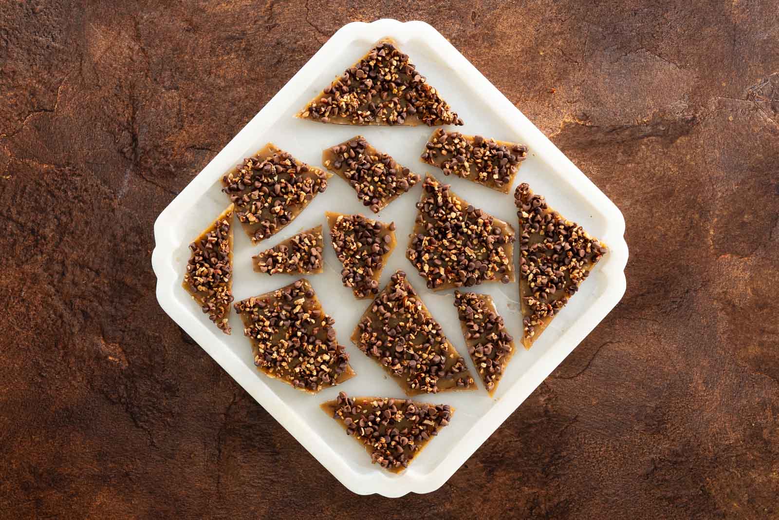 Overhead view of broken shards of English toffee topped with chocolate chips and pecans on parchment paper.