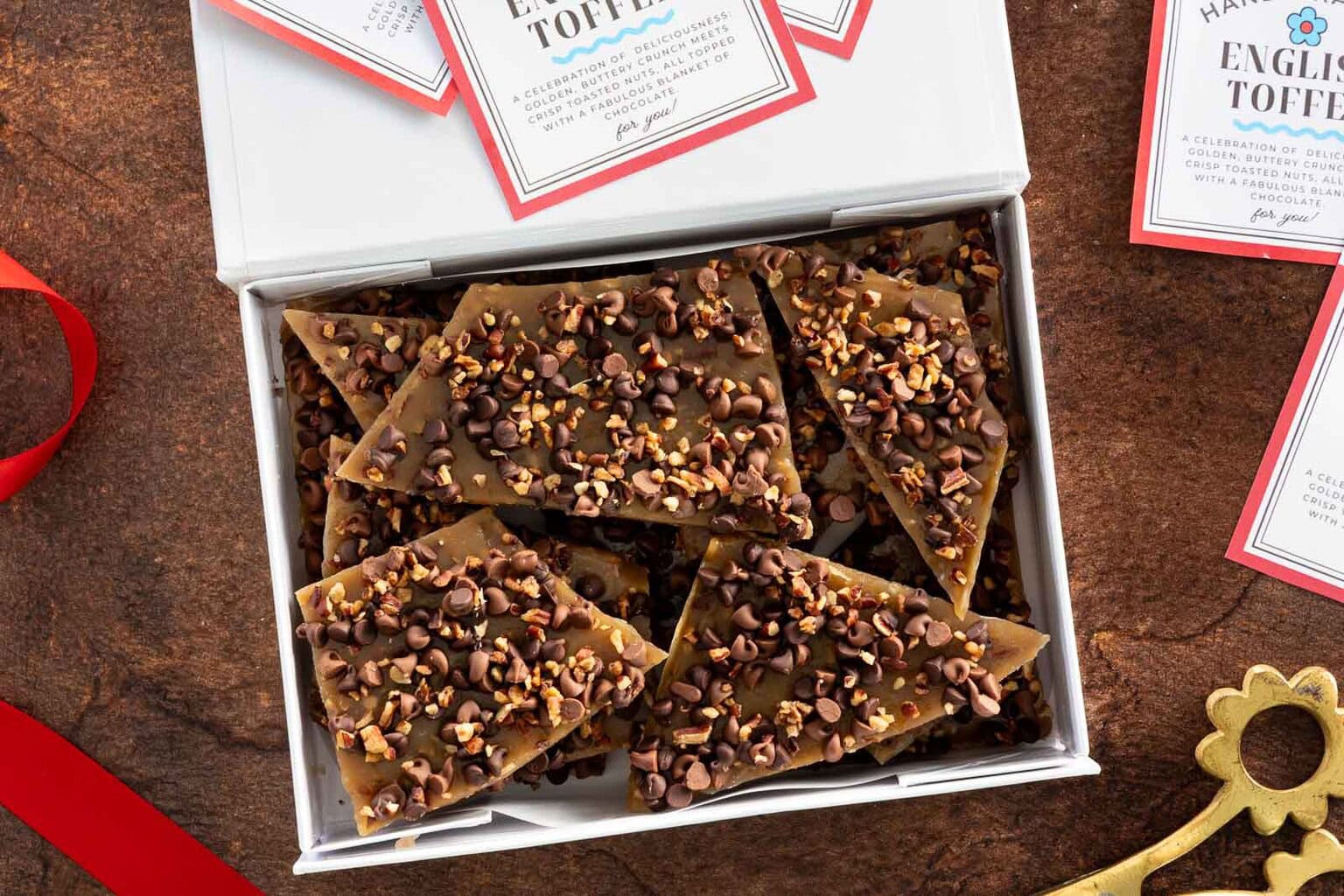 Close-up overhead shot of English toffee pieces in a white gift box with printable labels nearby.