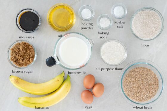Flat lay of labeled banana bran muffin ingredients on a marble countertop, including wheat bran, flour, eggs, bananas, buttermilk, oil, molasses, brown sugar, baking powder, baking soda, and kosher salt.