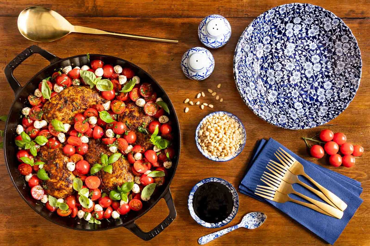 Cast iron skillet filled with golden-browned chicken thighs, cherry tomatoes, fresh basil, and mozzarella pearls on a wooden table with blue-and-white serving dishes.