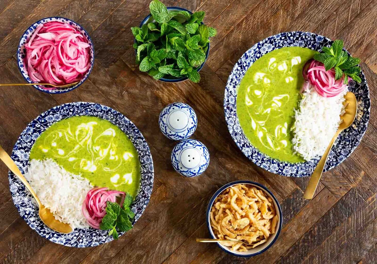 Bowls of vibrant green dahl served with white rice, pickled red onions, fresh mint, and crispy toppings on a wooden table.