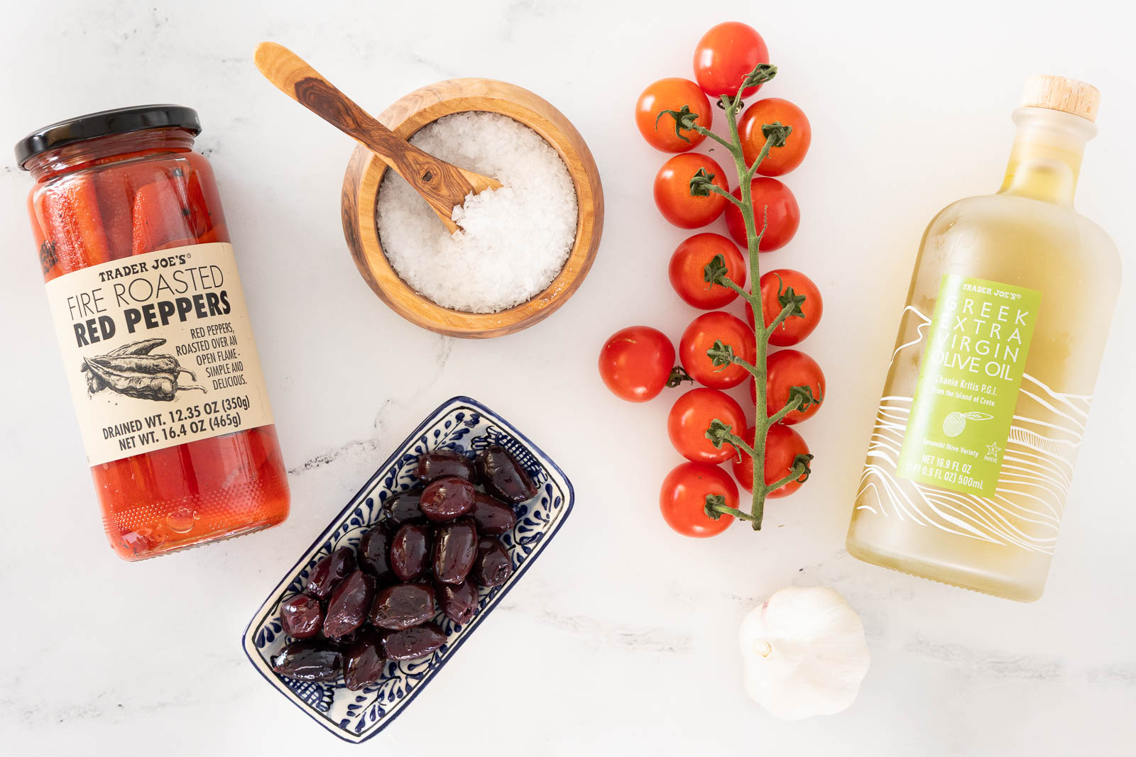 Overhead view of Mediterranean ingredients including roasted red peppers in a jar, cherry tomatoes on the vine, Kalamata olives, garlic, kosher salt, and olive oil arranged on a light surface.