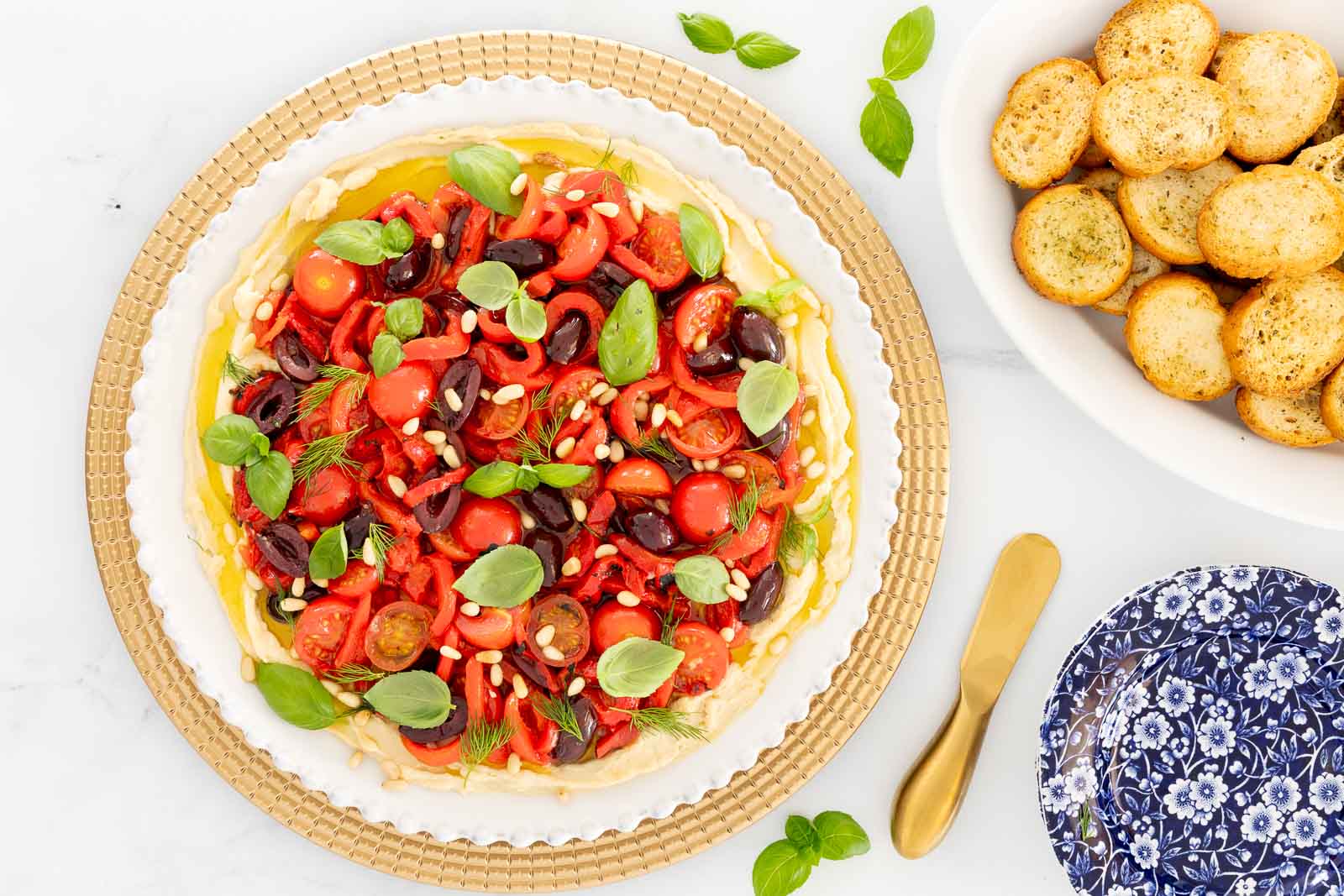 Overhead view of Italian white bean hummus topped with cherry tomatoes, roasted red peppers, Kalamata olives, pine nuts, basil, and fennel fronds, served with crostini nearby.