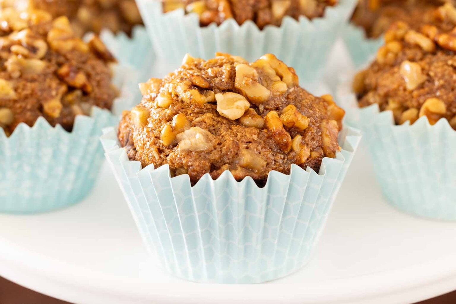 Close-up of a maple-glazed banana walnut bran muffin with a high domed top, baked in a pale blue liner and topped with chopped walnuts.