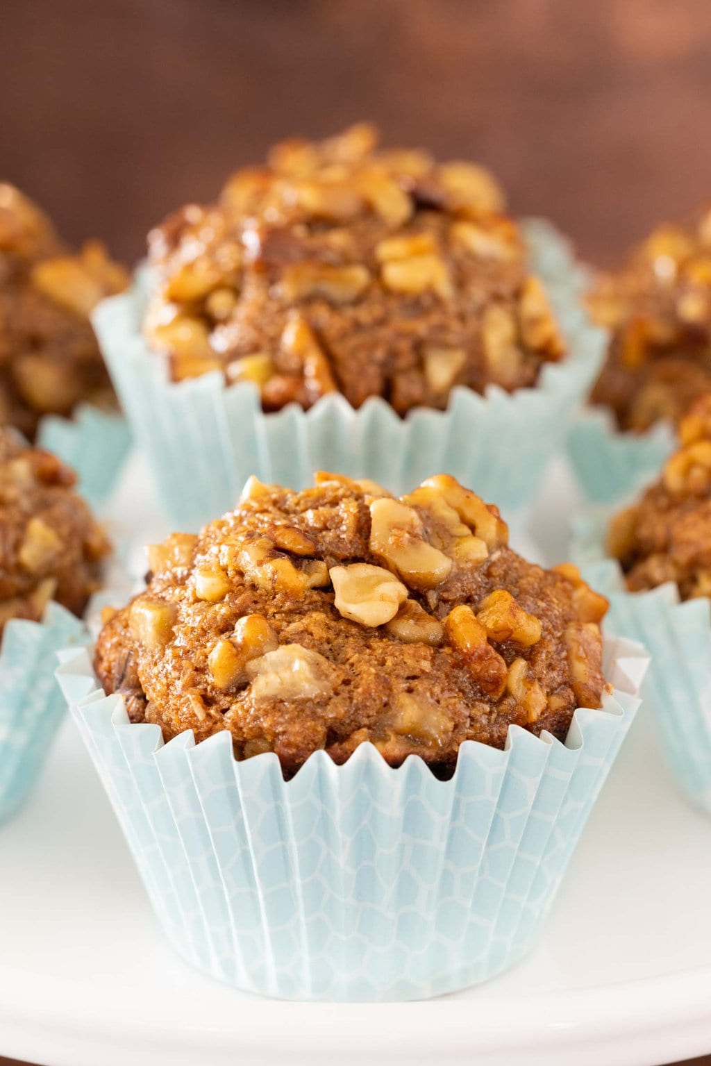 Close-up of a maple-glazed banana walnut bran muffin with a high domed top, baked in a pale blue liner and topped with chopped walnuts.