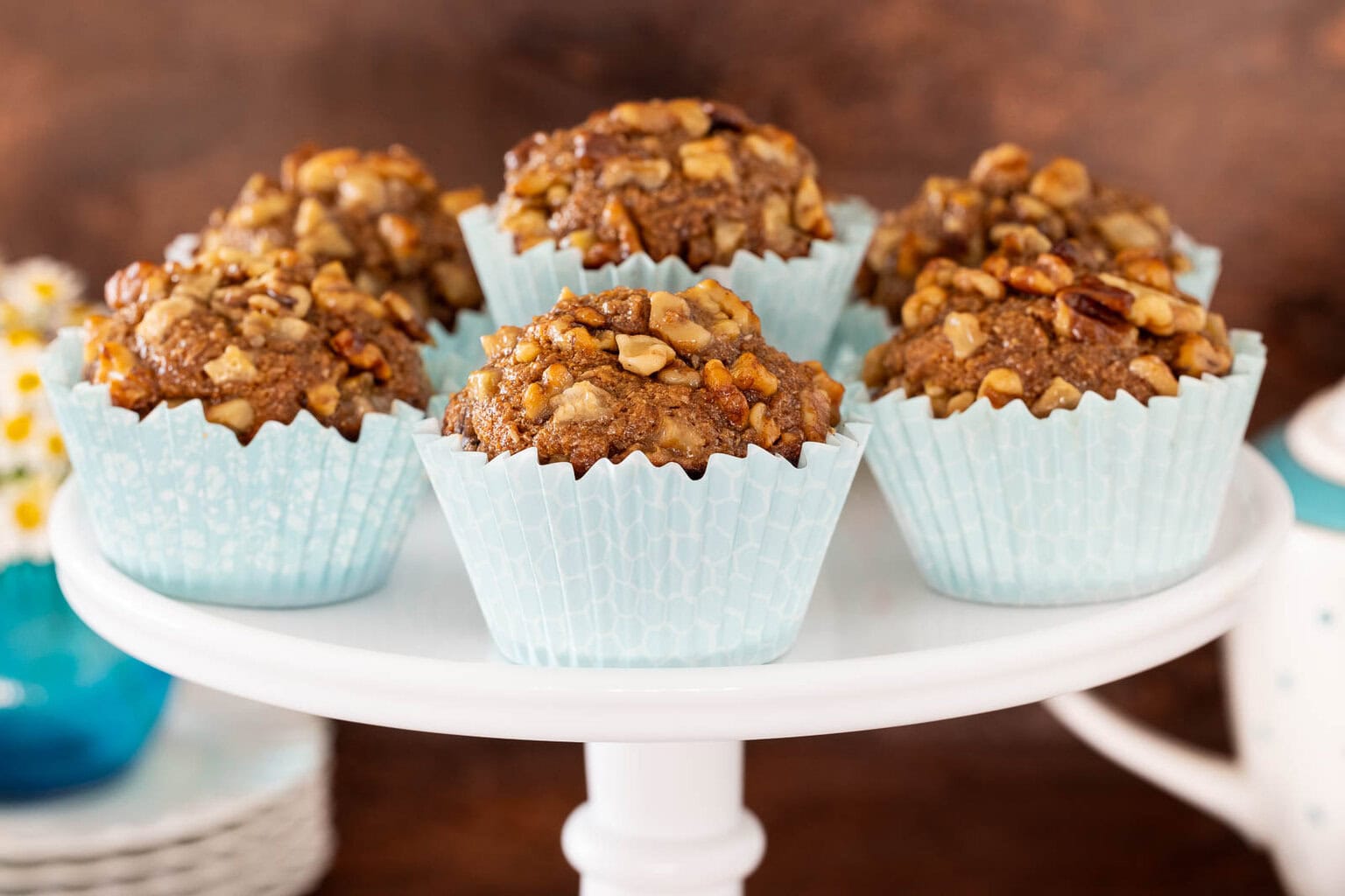 Maple-glazed banana walnut bran muffins with tall domed tops in pale blue liners, arranged on a white cake stand with a cozy kitchen background.
