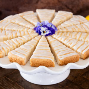 Lemon glazed shortbread wedges arranged on a white cake stand, drizzled with lemon icing and garnished with an edible flower.