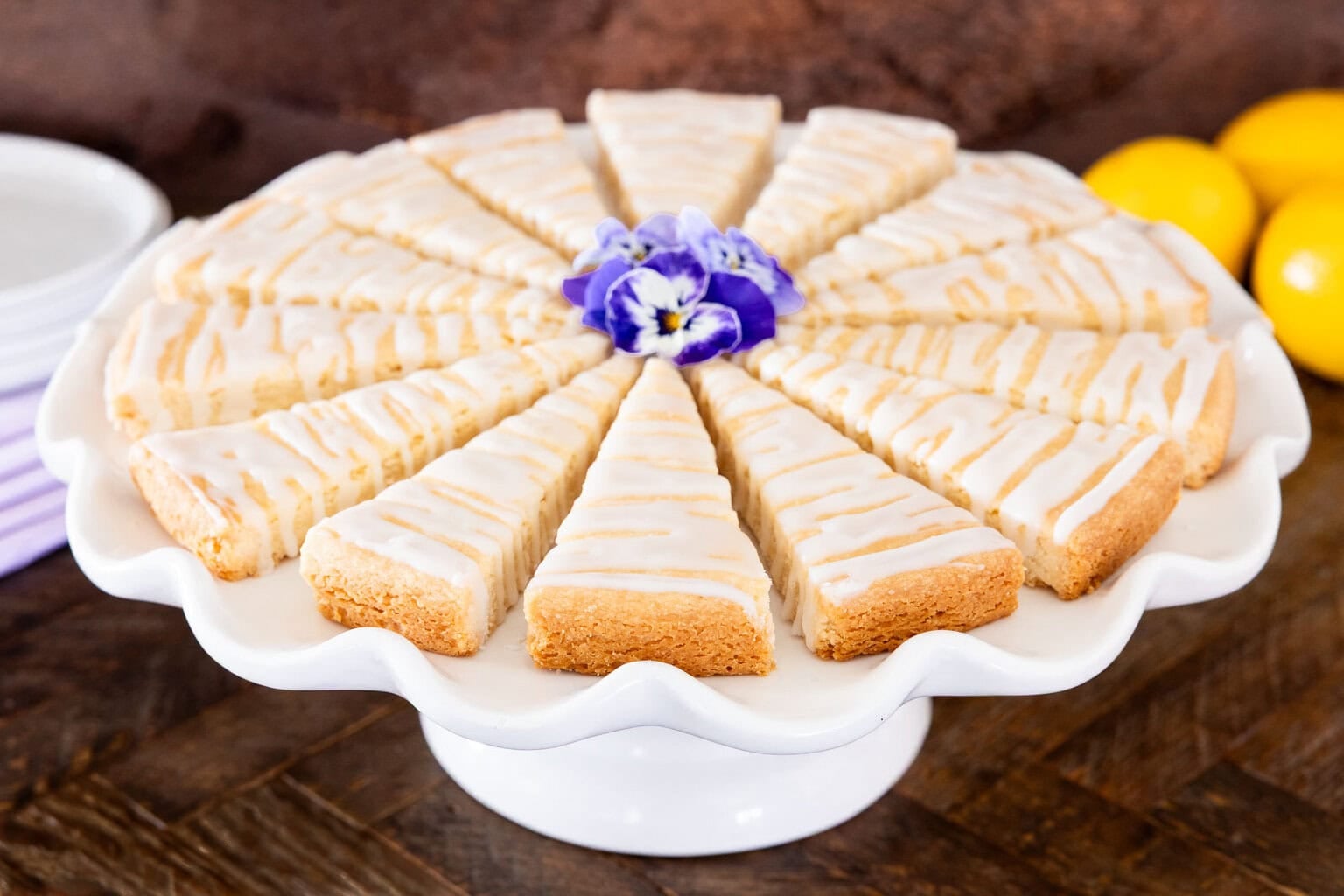 Lemon glazed shortbread wedges arranged on a white cake stand, drizzled with lemon icing and garnished with an edible flower.