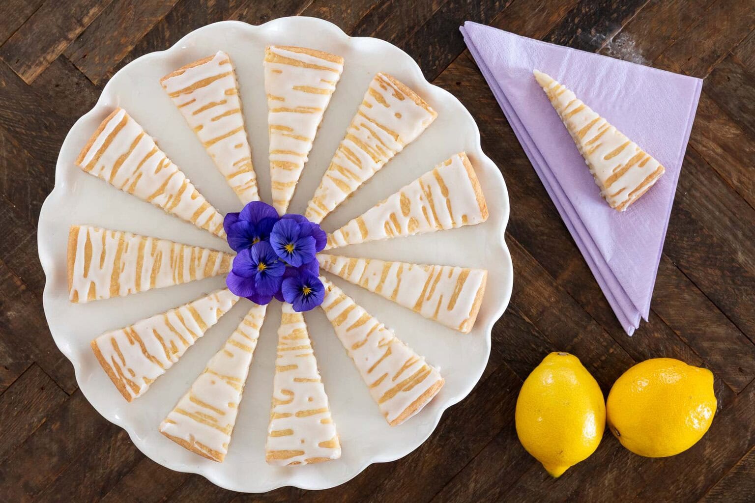 Slice of lemon-flavored cake with white icing and lemon drizzle, garnished with edible purple flowers.