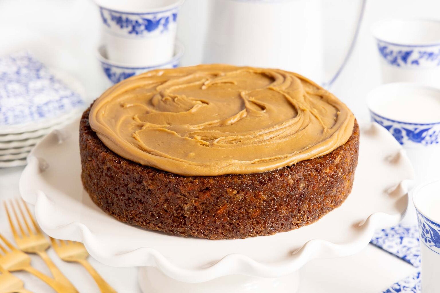 Single-layer carrot cake with brown sugar cream cheese frosting on a white scalloped cake stand, surrounded by blue and white dishes and gold forks.