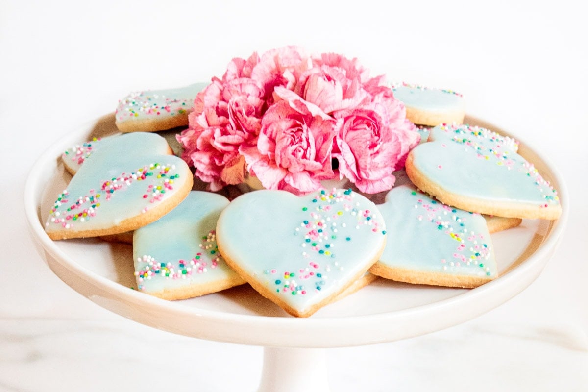 Heart-shaped Valentine shortbread cookies with pastel blue icing and colorful sprinkles arranged on a white cake stand with pink flowers.