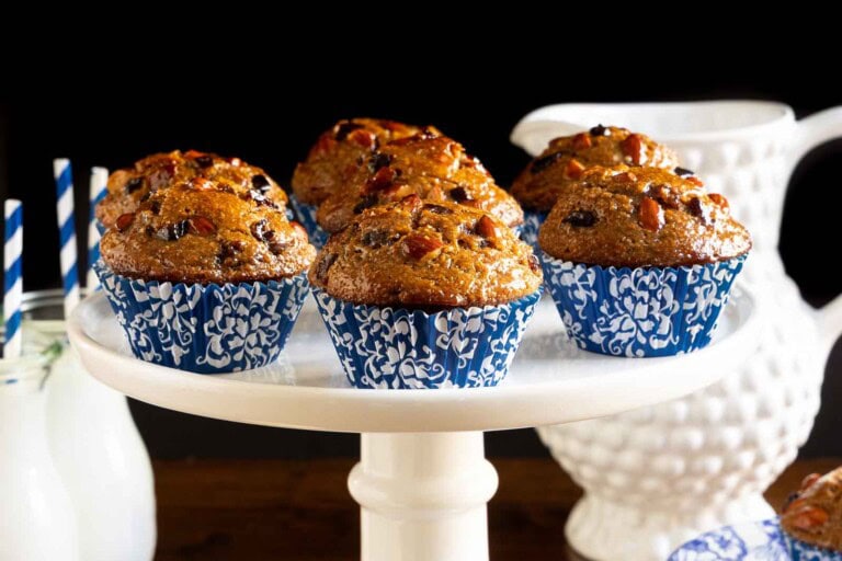 Honey glazed almond cherry bran muffins topped with sliced almonds and dried cherries in blue patterned liners on a white cake stand.