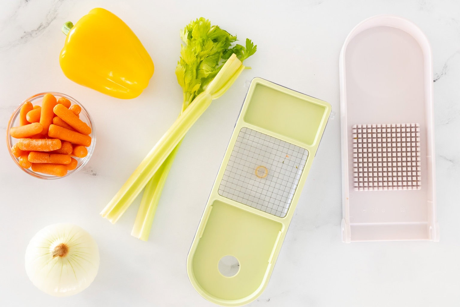 Flat lay of vegetables for Italian Grandmother Minestrone including onion, celery, carrots, and yellow bell pepper, styled with a vegetable chopper on a white countertop.
