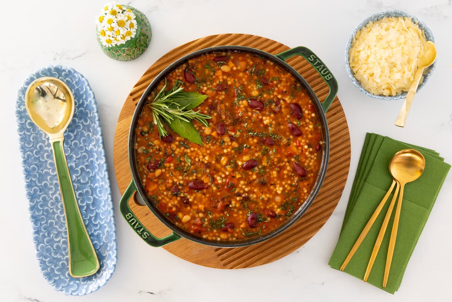 Italian Grandmother Minestrone soup with beans, vegetables, and acini de pepe pasta in a green Staub Dutch oven, styled overhead with Parmesan cheese, green napkins, and gold serving spoons.
