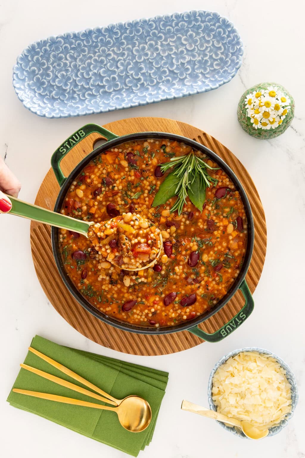 Italian Grandmother Minestrone soup with beans, vegetables, and acini de pepe pasta in a basil green Staub Dutch oven, styled overhead with Parmesan cheese, green napkins, and a serving spoon.