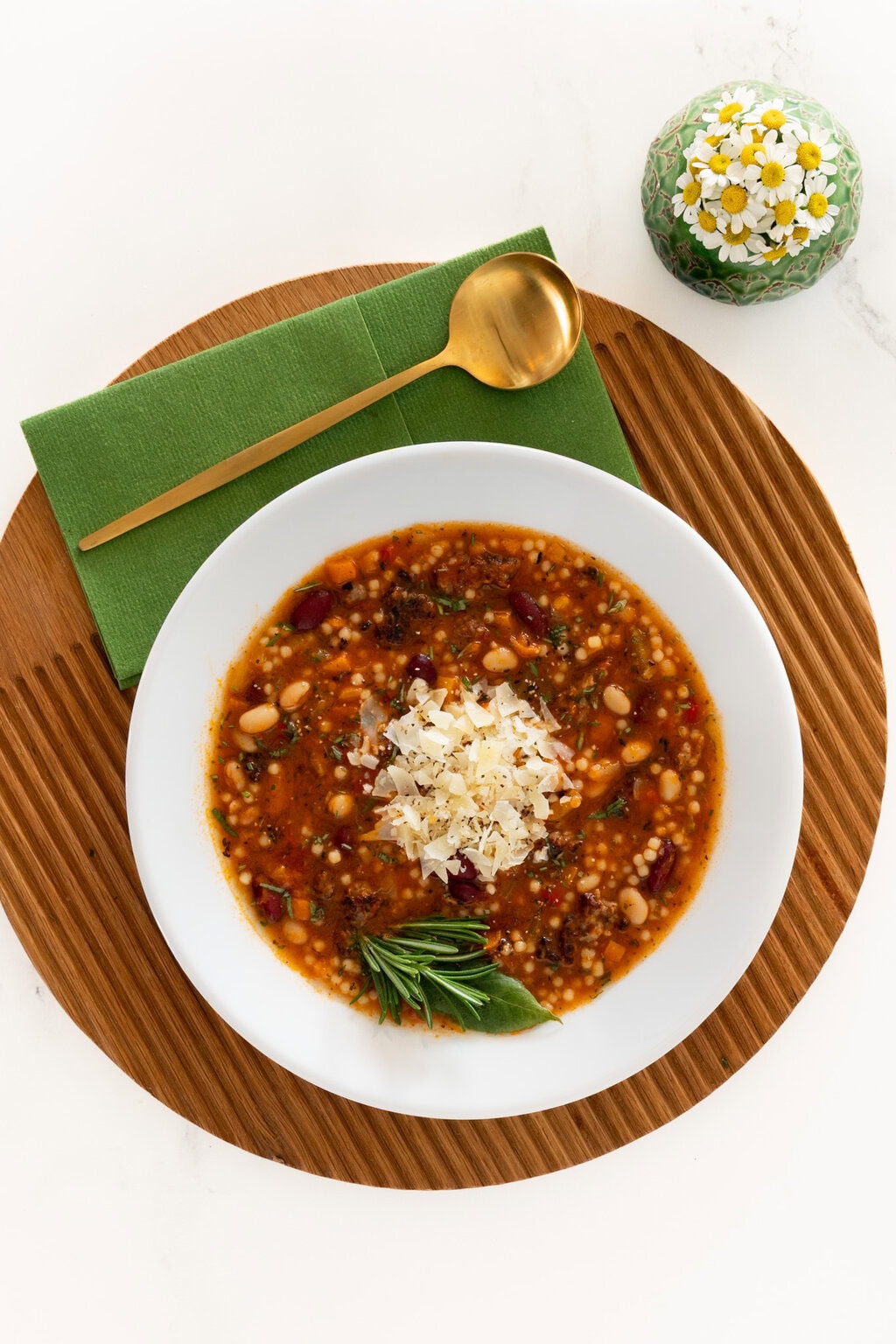 Bowl of Italian Grandmother Minestrone soup with beans, vegetables, acini de pepe pasta, grated Parmesan, and fresh rosemary, styled overhead on a wooden board with green napkin and gold spoon.
