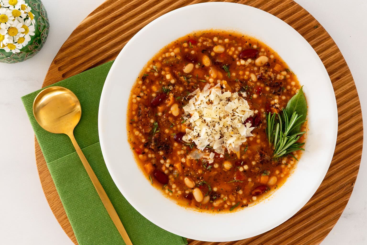 Close-up overhead view of Italian Grandmother Minestrone soup with beans, vegetables, acini de pepe pasta, grated Parmesan, and rosemary, served in a white bowl on a wooden board.