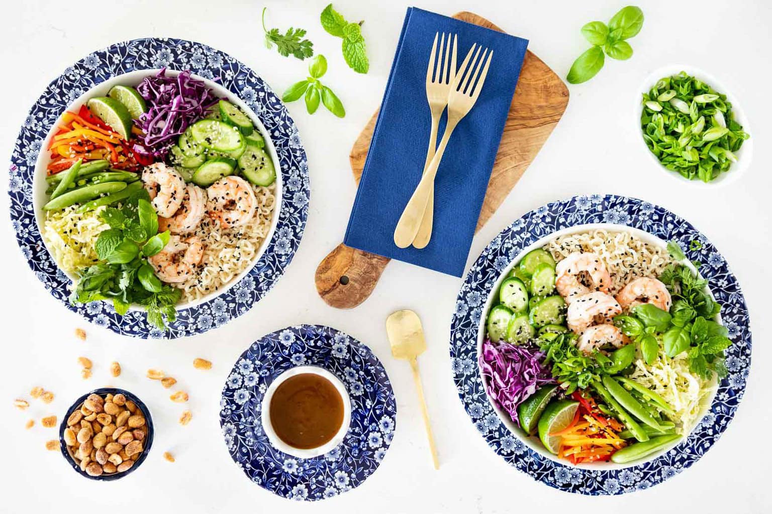 Two bowls of Asian noodle salad with shrimp, cabbage, cucumbers, carrots, fresh herbs, and peanut dressing on a white background.