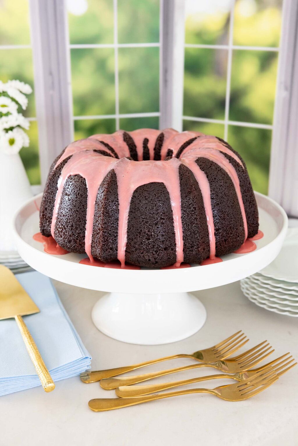 Chocolate pound cake with a thinner fresh strawberry icing dripping down the sides on a white cake stand.