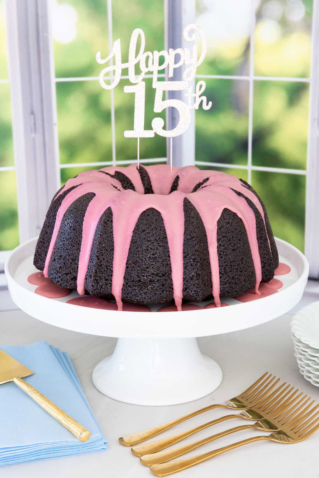 Chocolate pound cake with strawberry icing and a Happy 15th cake topper on a white cake stand.