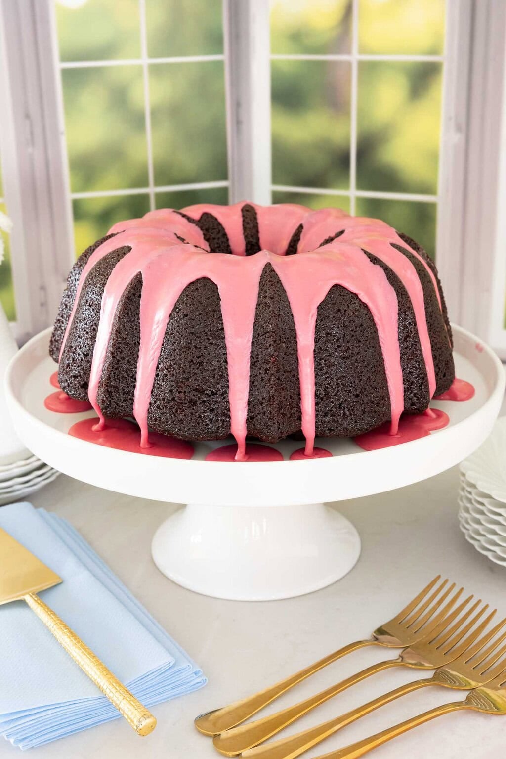 Chocolate pound cake with fresh strawberry icing dripping down the sides on a white cake stand with gold forks and blue napkins.