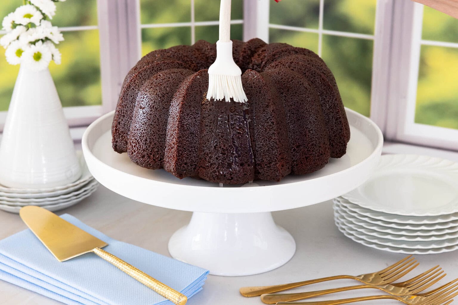 Brushing glaze onto warm chocolate pound cake on a white cake stand.