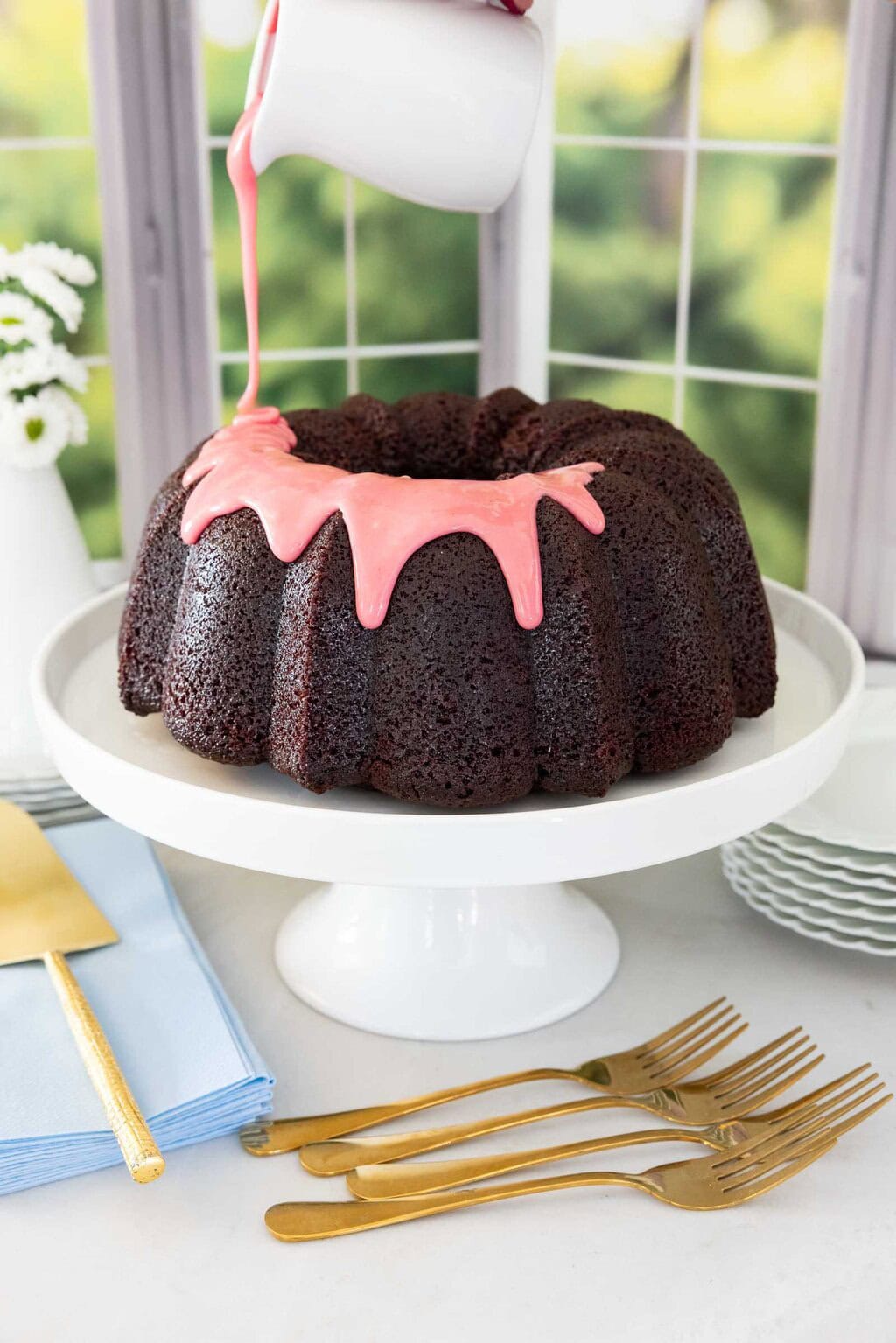 Pouring fresh strawberry icing over chocolate pound cake on a white cake stand.
