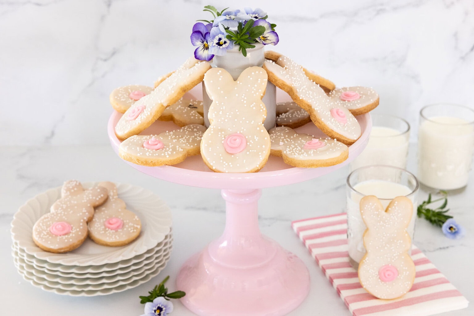 Easter bunny shortbread cookies with glaze and pink tails displayed on a pink cake stand with spring flowers and milk