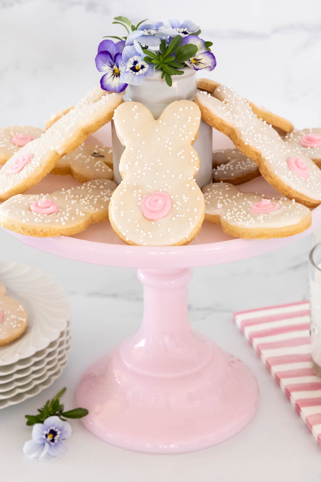 Easter bunny shortbread cookies with white glaze and pink buttercream tails displayed on a pink cake stand with flowers