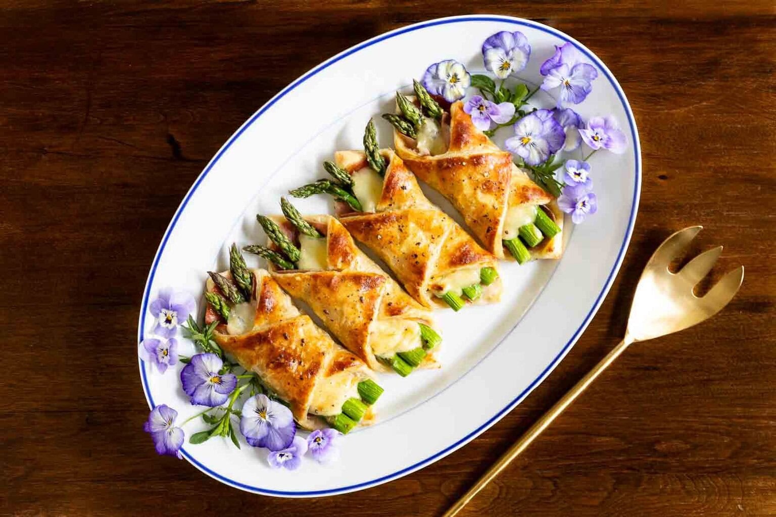 Golden puff pastry bundles filled with asparagus and cheese on a white platter garnished with purple edible flowers.
