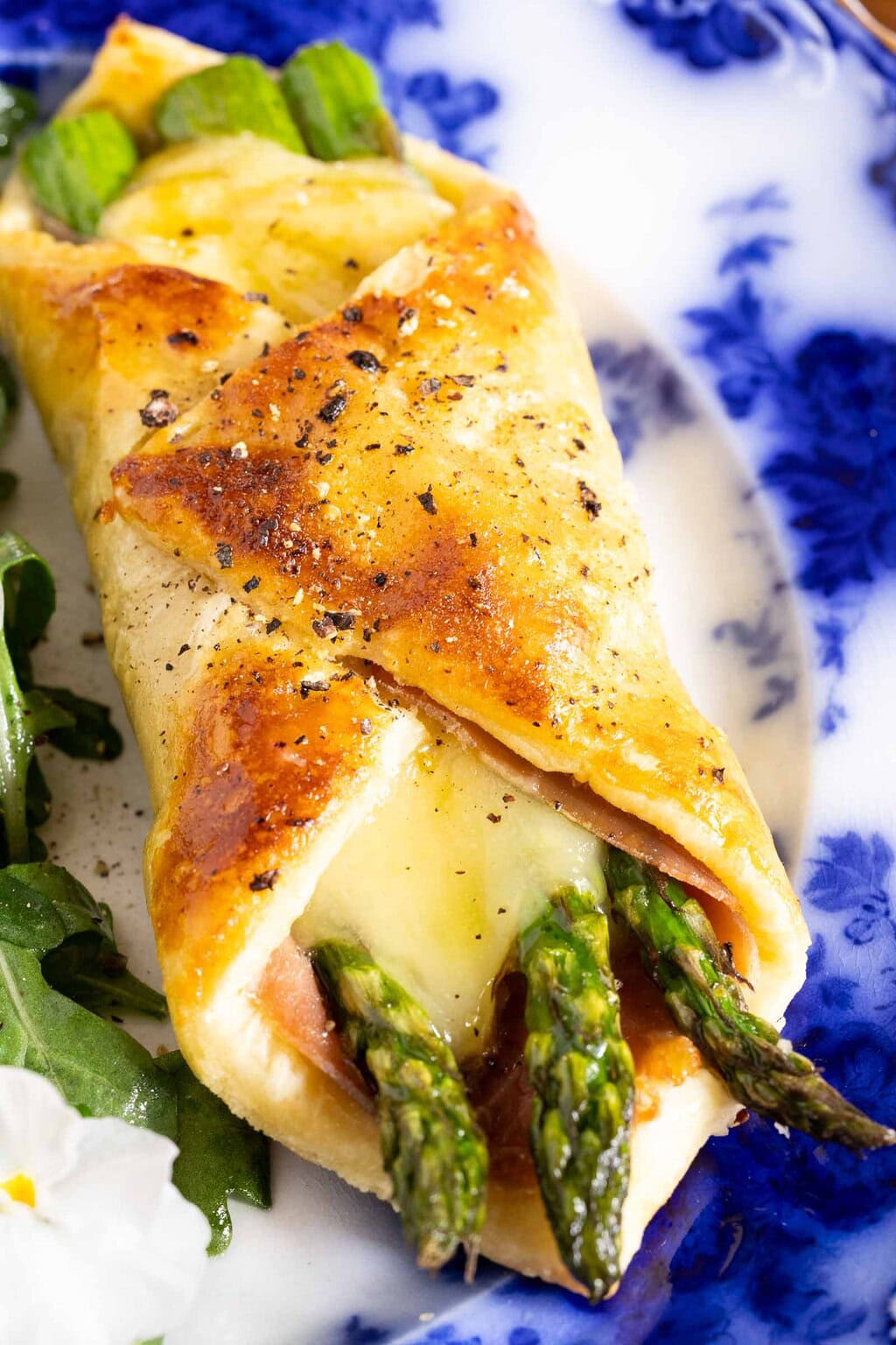 Close-up of golden puff pastry filled with asparagus, ham and melted cheese on a blue and white plate
.