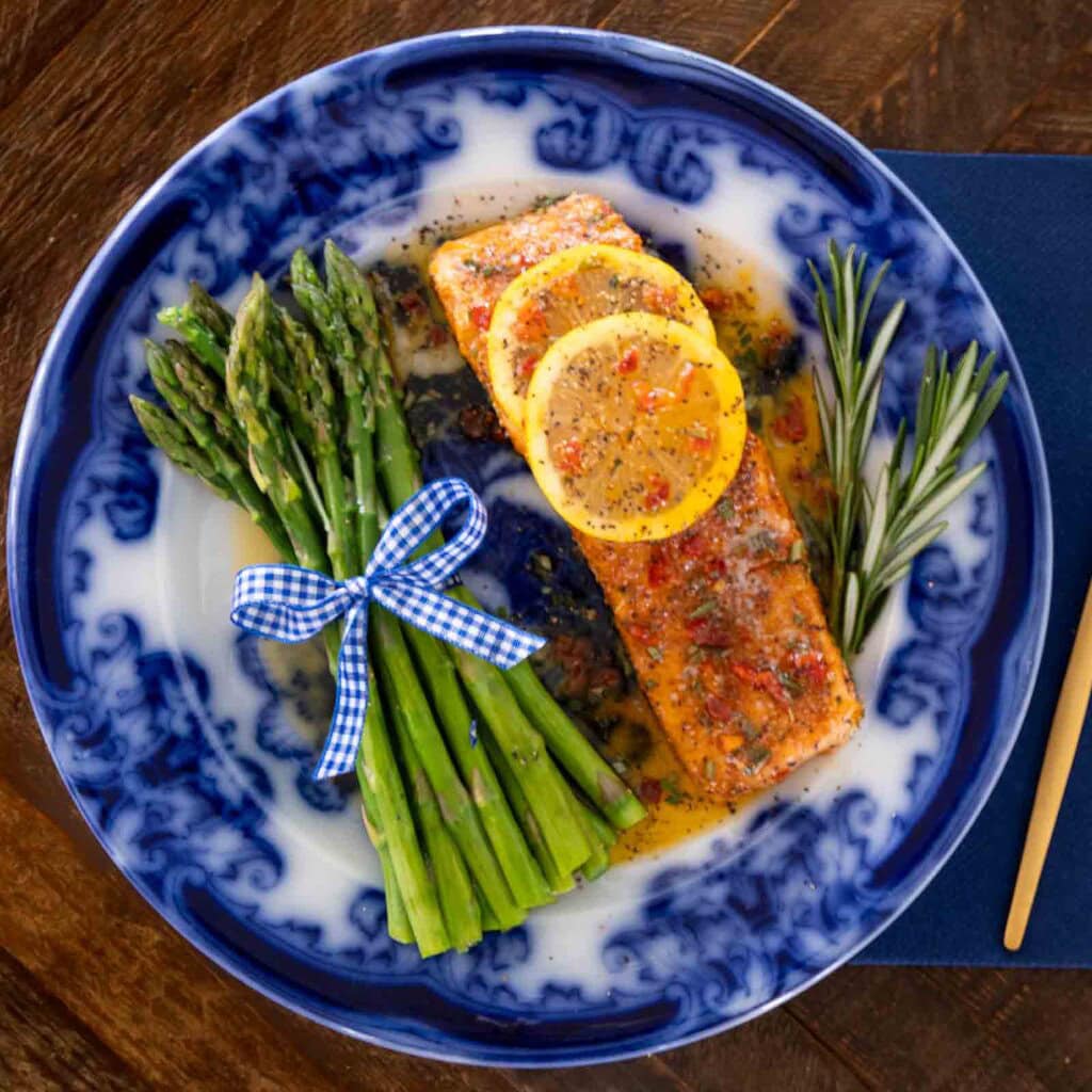 Pan-seared salmon with garlic herb butter, lemon slices and sun-dried tomatoes served with asparagus on a blue and whiteplate.