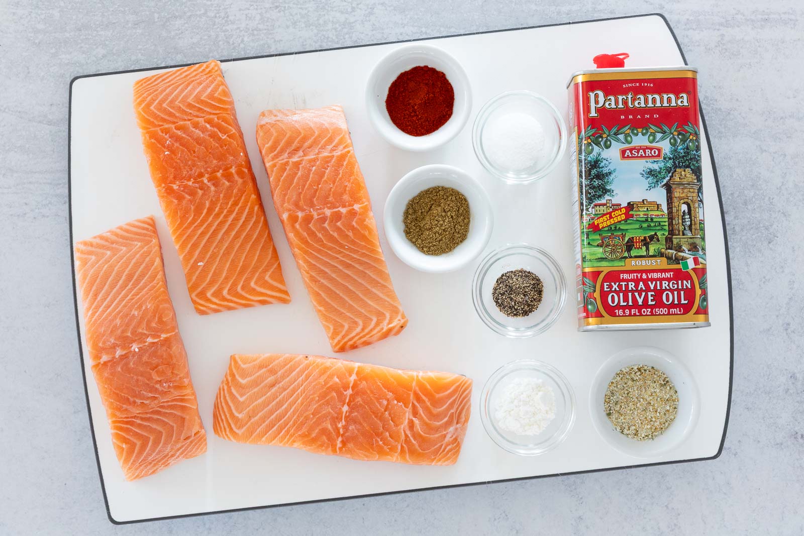 Fresh salmon fillets with small bowls of spices and olive oil arranged on a cutting board for Italian garlic butter pan-seared salmon.