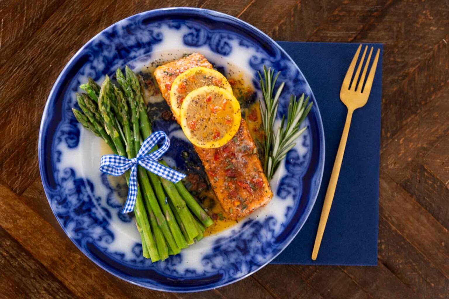 Pan-seared salmon with garlic herb butter, lemon slices and sun-dried tomatoes served with asparagus on a blue plate.