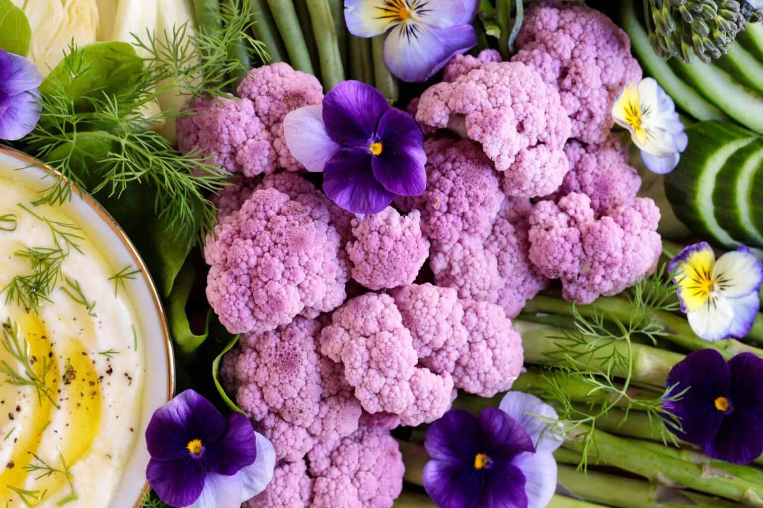 Close-up of spring vegetable platter with purple cauliflower, asparagus, cucumbers, snap peas and creamy lemon feta dip garnished with herbs and edible flowers